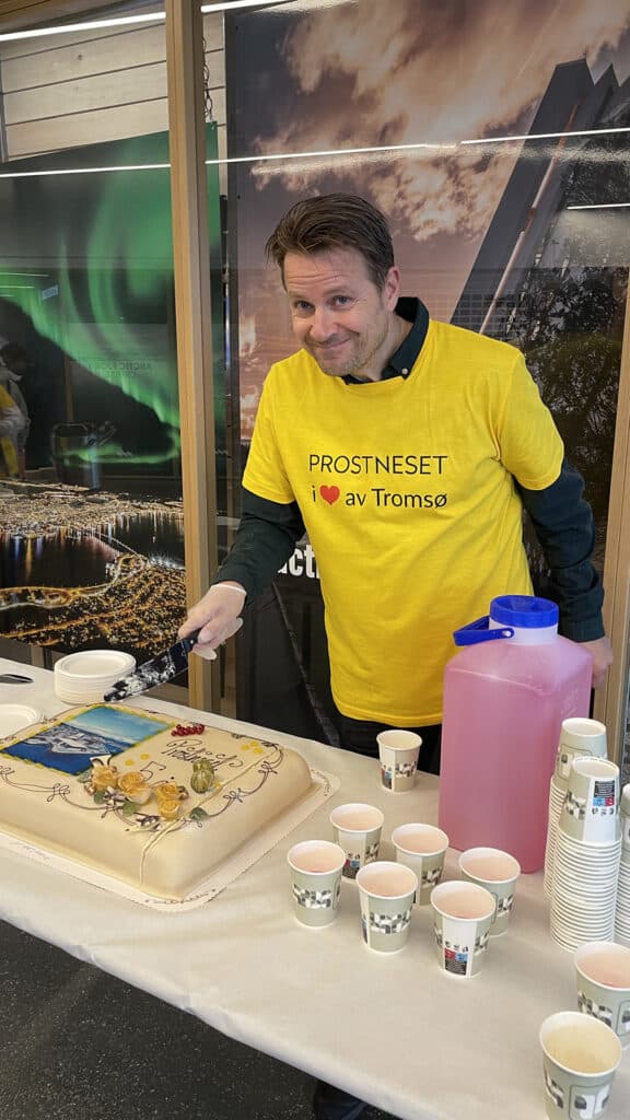 En mann i en gul PROSTNESET I ♥ av Tromsø-skjorte smiler mens han serverer en pyntet kake ved et bord med pappkrus og en mugge med rosa drikke. Bakgrunnen viser naturskjønne bilder av Tromsø.