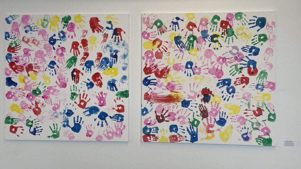 To lerret på en hvit vegg, hvert dekket av fargerike håndavtrykk i forskjellige størrelser og farger, inkludert rød, blå, grønn, gul og rosa, noe som skaper en levende og leken utstilling.