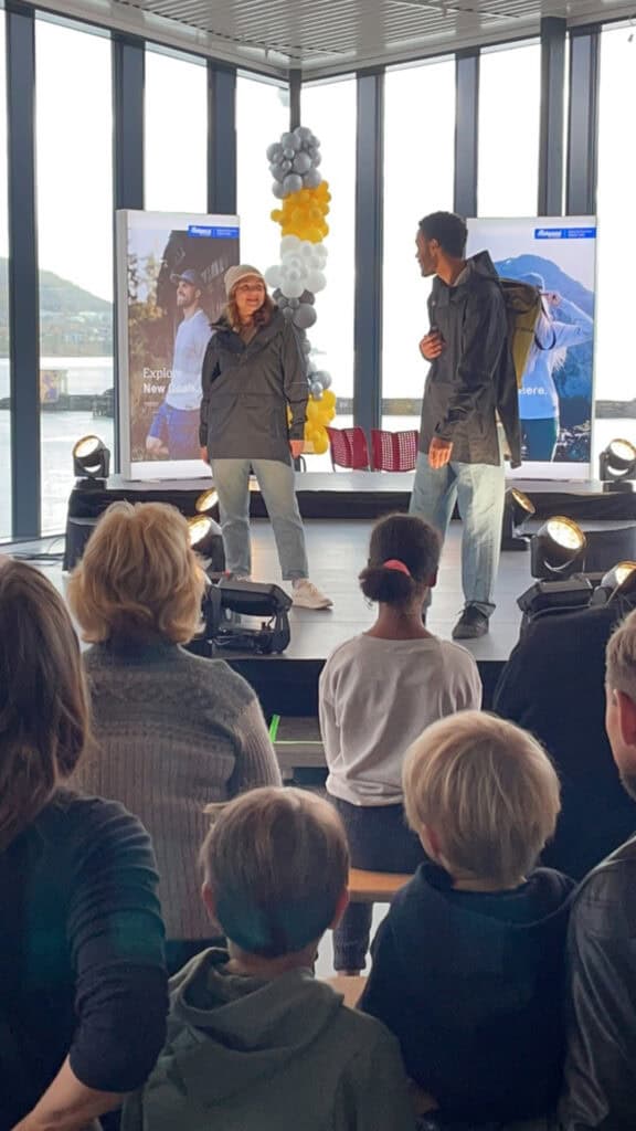 To personer i jakker og jeans står på en scene, vendt mot hverandre, med et publikum som ser på. Bak dem er det plakater, ballonger og store vinduer som viser en utendørsscene. Scenelys lyser opp scenen.