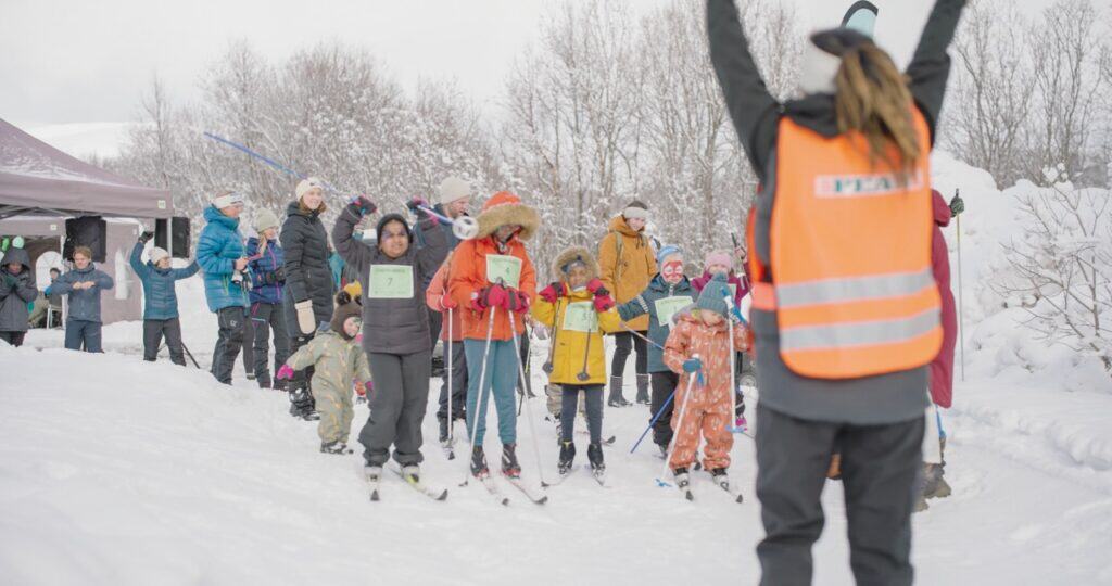 En gruppe barn i vinterklær står på ski ved en snødekt startstrek og holder sertifikater. En voksen i en knalloransje vest står i forgrunnen med armene hevet, mens tilskuere ser på fra siden.