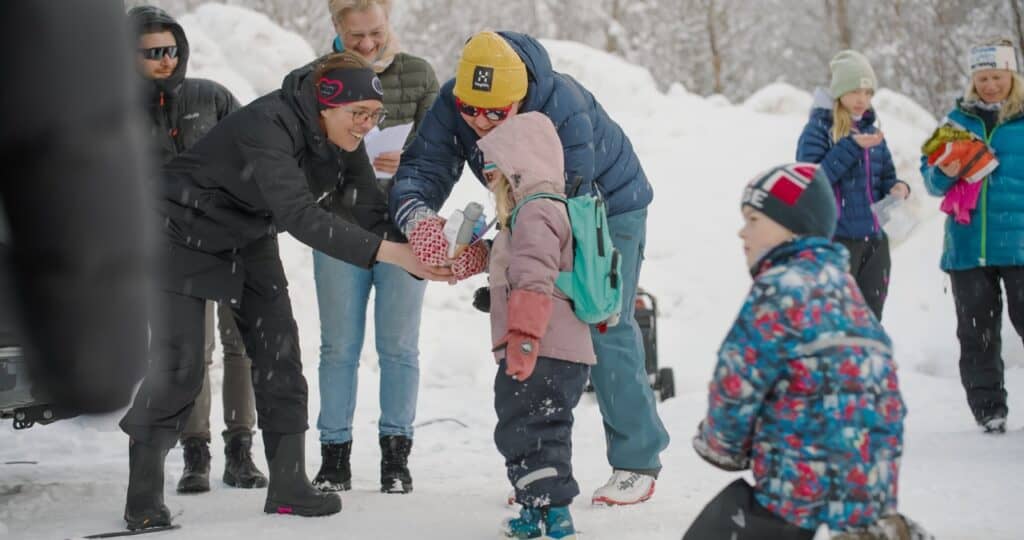 En gruppe voksne og barn kledd i vinterklær samles utendørs i snøen. To voksne gir en gave til et barn iført rosa jakke og blågrønn ryggsekk. Andre ser på og smiler i den snødekte bakgrunnen.