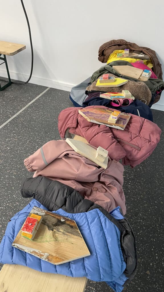 Flere fargerike jakker henger over en benk, hver med et trebrett som inneholder en juiceboks og en pakket snacks på toppen. Omgivelsene ser ut til å være innendørs med et flekkete gulv og en hvit vegg.