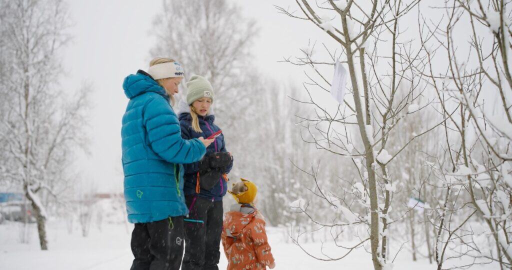 Tre personer, kledd i vinterklær, står i nærheten av nakne trær dekket av snø. En voksen holder en telefon, mens en annen voksen og et barn ser opp på grenene. Snø dekker bakken og trærne i bakgrunnen.