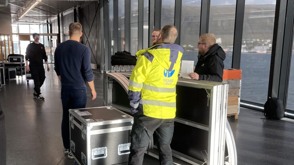 Fire menn står rundt metallkasser og utstyr ved store vinduer med utsikt over vannet. Én har på seg en knallgul Technical Crew-jakke, mens andre er kledd i mørke klær og ser ut til å diskutere oppsettsplaner.