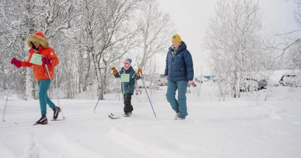 Tre personer, inkludert et barn, står på ski sammen på en snødekt løype blant bladløse trær. Alle er kledd i vinterklær og ser ut til å nyte det kalde utendørsmiljøet.