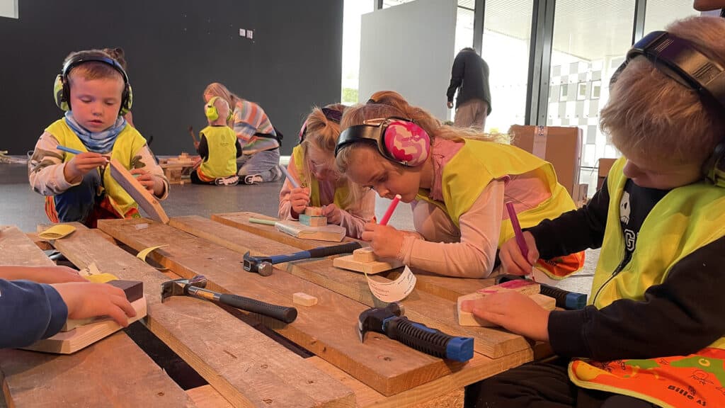Barn iført sikkerhetsvester og hodetelefoner bruker tusjer og verktøy til å jobbe med treklosser ved et bord. Omgivelsene ser ut til å være et klasserom eller verksted, med voksne som fører tilsyn i bakgrunnen.