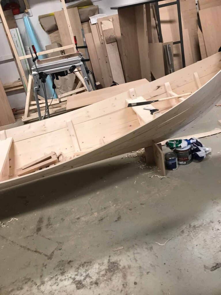 En trebåt under bygging i et verksted, omgitt av verktøy, trebiter og bokser med forsyninger. Båtrammen er delvis montert og står på et betonggulv.