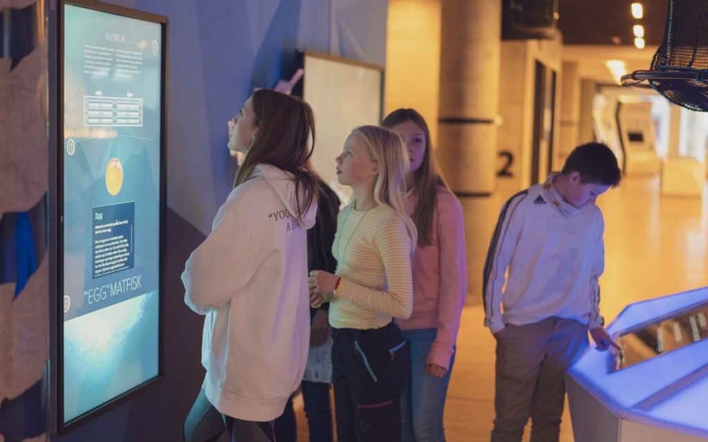 En gruppe barn står innendørs og samhandler med en stor digital skjerm og et berøringsbord i det som ser ut til å være en utstilling på et vitenskaps- eller teknologimuseum.