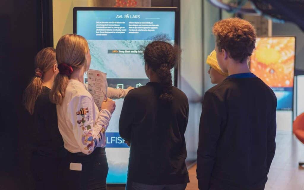 Fire barn står foran en interaktiv skjerm på et museum eller vitenskapssenter, leser informasjon og peker på skjermen sammen. Ett barn holder et arbeidsark.