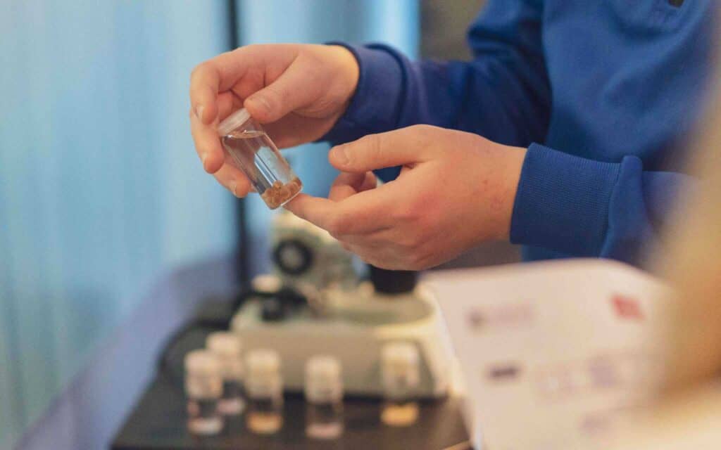 En person i blå skjorte holder et lite glass med brunlige prøver inni, med et mikroskop og andre glass uskarpe i bakgrunnen, og et dokument delvis synlig i forgrunnen.