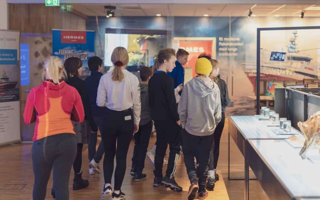 En gruppe unge mennesker, for det meste kledd i uformelt sportstøy, går sammen gjennom et sterkt opplyst utstillingsområde med utstillinger og informasjonsskilt på veggene.