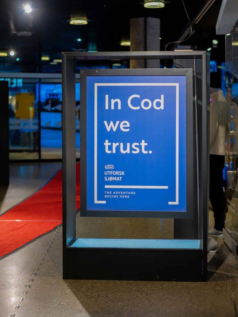 Et blått skilt med hvit tekst som «In Cod we trust» står innendørs på et polert gulv ved siden av en rød teppe. Logoen til Utforsk Sjømat er synlig nederst på skiltet.